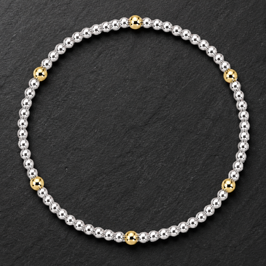 A beaded stretch bracelet featuring a mix of polished silver and yellow gold beads, arranged in a circular shape on a dark textured slate background.