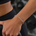 A close-up of a person's wrist wearing a delicate beaded bracelet with alternating silver and gold beads, set against a blurred gym background.