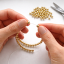 Close-up of hands threading gold beads onto a string, showing the handmade jewelry-making process-desktop